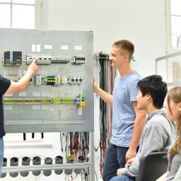 3 Auszubildende und der Ausbilder stehen vor einer Schalttafel 