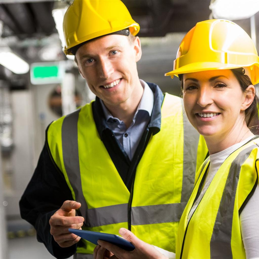 2 Mechaniker, ein junger Mann und eine junge Frau, in gelben Warnwesten, lächeln freundlich in die Kamera